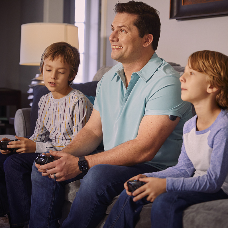 Man playing video games with his kids while wearing an implantable CGM on his arm.