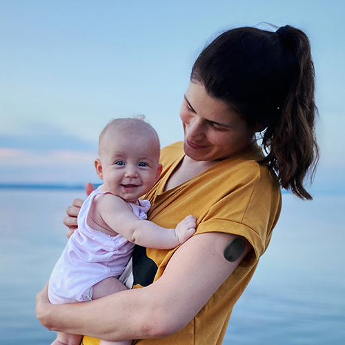 Woman wearing an Eversense long term CGM holds her baby on the beach.