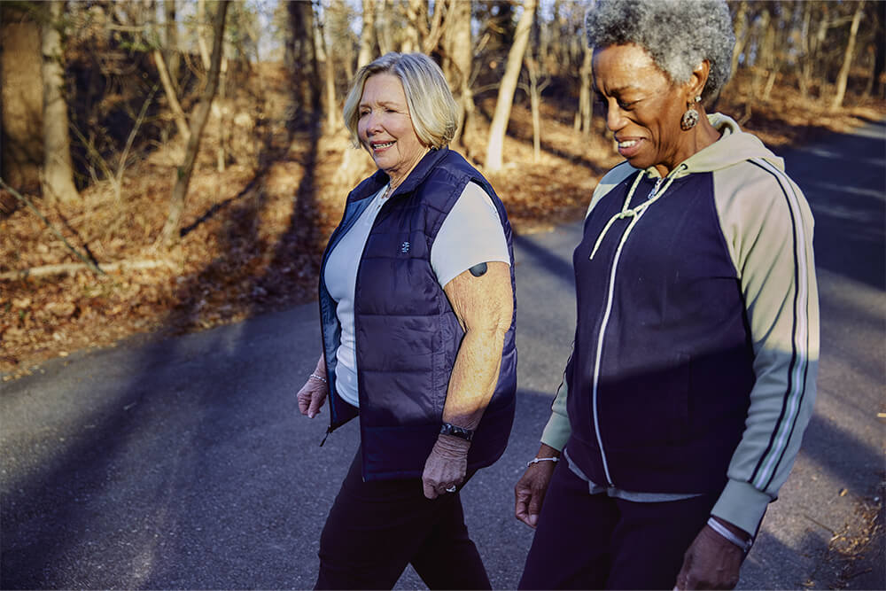 Two older women walking and discussing continuous glucose monitor insurance coverage.