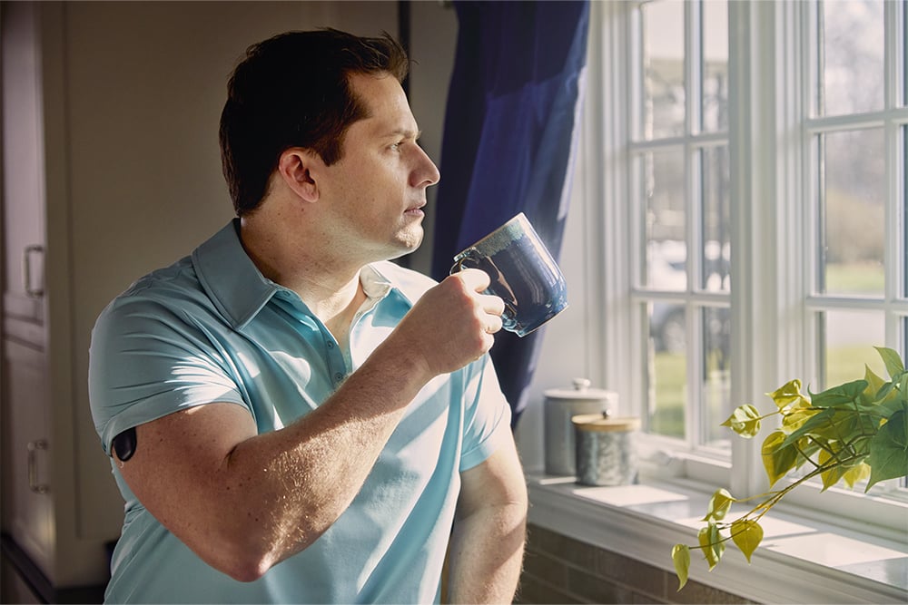 Man peering out of the window while wearing a CGM device. 
